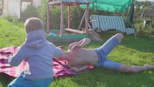 Två barn leka på gräsmattan på en varm sommardag. Barn skratta, de kör och faller på gräsmattan. Underhållning utomhus — Stockvideo
