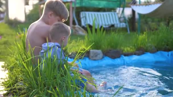 Twee kinderen spelen in de buurt van een meertje, bloemen en gras groeien rond het meer. Kinderen maken spatten van water met hun voeten. Warme zomerdag. Gelukkige jeugd — Stockvideo