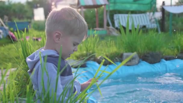 Le garçon est assis près d'un petit lac. L'enfant crée des éclaboussures d'eau avec ses pieds. Une chaude journée d'été. Bonne enfance — Video