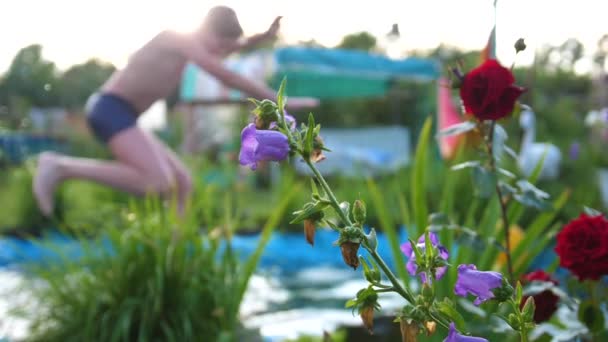 Los niños nadan en un pequeño lago en un caluroso día de verano. El chico salta al agua, crea salpicaduras de agua. Jardín, flores y plantas alrededor del lago. Feliz infancia. — Vídeo de stock