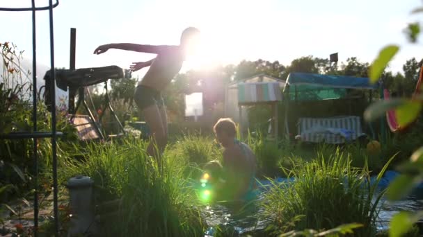 La familia nada en un pequeño lago en un caluroso día de verano. El chico salta al agua, crea salpicaduras de agua. Jardín, flores y plantas alrededor del lago. Feliz infancia. — Vídeos de Stock