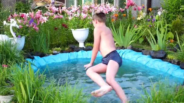 Il ragazzo sta nuotando in un piccolo lago in una calda giornata estiva. Il ragazzo salta in acqua, crea spruzzi d'acqua. Giardino, fiori e piante intorno al lago. Buona infanzia. — Video Stock