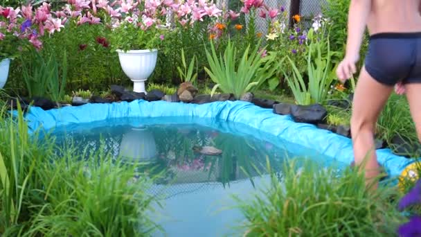 Les enfants nagent dans un petit lac par une chaude journée d'été. Le garçon saute dans l'eau, crée des éclaboussures d'eau. Jardin, fleurs et plantes autour du lac. Bonne enfance — Video