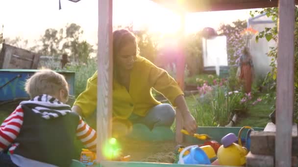 Una madre joven con un niño jugando en la caja de arena. Día de verano, rayos de sol en el parque infantil — Vídeos de Stock