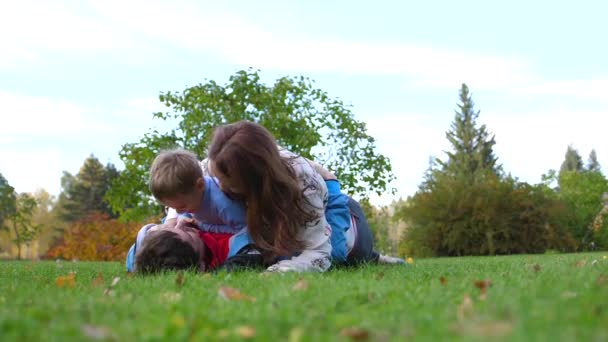 Familia feliz tendida en el césped. Madre y padre jugando con su hijo, el niño sonríe. Sobre el fondo del sol y del cielo, rostros felices de padres e hijos . — Vídeos de Stock