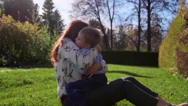 Щаслива сім'я відпочиває на газоні. Мама з ніжністю і любов'ю обіймає свою дитину, син сміється. Щасливе дитинство — стокове відео