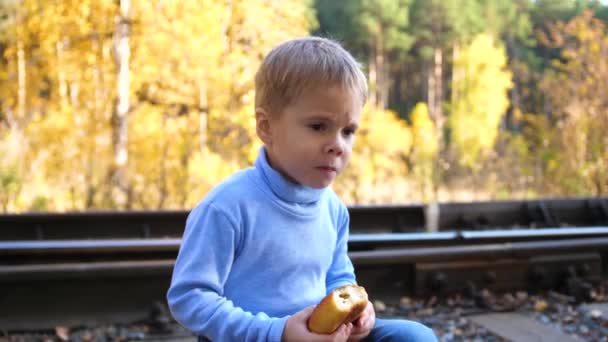 El chico se sienta y se come un bollo en las vías del tren. Otoño día cálido . — Vídeos de Stock