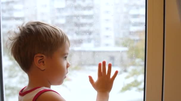 Un bambino sta vicino a una finestra e guarda la neve cadere per strada. La mano di primo piano sulla finestra di vetro — Video Stock