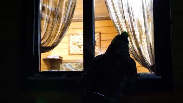 Noche de invierno. En una habitación cálida y luminosa cerca de la ventana hay un hombre joven y llama a la ventana. Disparos al aire libre . — Vídeos de Stock