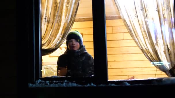 Noche de invierno. En una habitación cálida y luminosa cerca de la ventana se encuentra un hombre joven. lanzando una bola de nieve en la ventana — Vídeo de stock