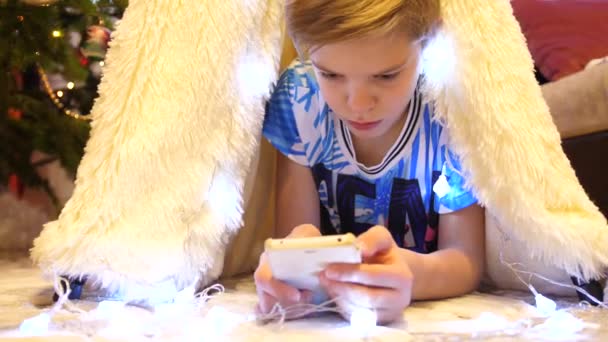 Ο έφηβος παίζει με το smartphone στο παιδικό δωμάτιο σε μια σκηνή με ένα ελαφρύ Χριστούγεννα. Ευτυχισμένη παιδική ηλικία — Αρχείο Βίντεο