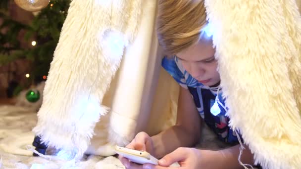 L'adolescent joue sur le smartphone dans la chambre des enfants dans une tente avec une lumière de Noël. Bonne enfance — Video