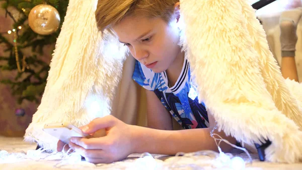 Ο έφηβος παίζει με το smartphone στο παιδικό δωμάτιο σε μια σκηνή με ένα ελαφρύ Χριστούγεννα. Ευτυχισμένη παιδική ηλικία — Φωτογραφία Αρχείου
