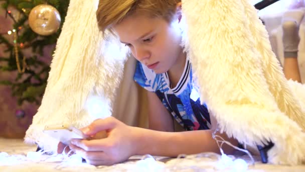 Der Teenager spielt mit dem Smartphone im Kinderzimmer in einem Zelt mit Weihnachtsbeleuchtung. Glückliche Kindheit — Stockvideo