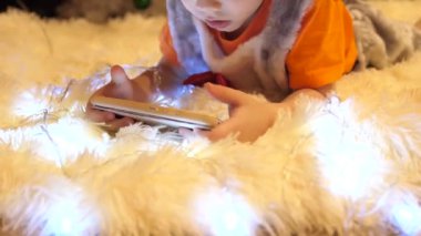 Çocuk yumuşak beyaz battaniye çocuk odasında yatıyor. Smartphone çizgi film izliyor. Noel çelenk. Mutlu çocukluk