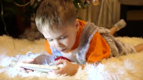 Το παιδί βρίσκεται σε ένα μαλακό κάλυμμα λευκό στο παιδικό δωμάτιο. Αυτός είναι βλέποντας κινούμενα σχέδια στο smartphone. Χριστουγεννιάτικη γιρλάντα. Ευτυχισμένη παιδική ηλικία — Αρχείο Βίντεο