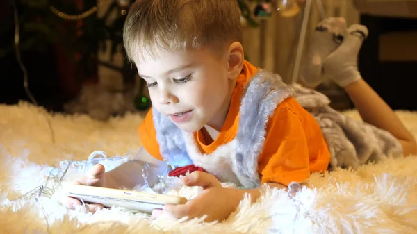 L'enfant repose sur une couverture blanche douce dans la chambre des enfants. Il regarde des dessins animés sur le smartphone. Guirlande de Noël. Bonne enfance Image En Vente