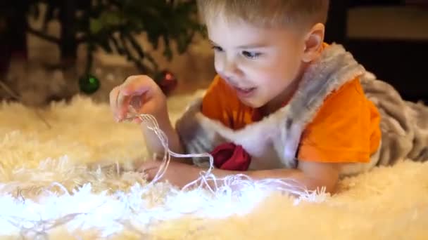 El niño juega en la habitación de los niños con una luz de Navidad, una guirnalda. Feliz infancia. — Vídeos de Stock