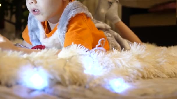 Il bambino giace su una morbida coperta bianca nella stanza dei bambini. Sta guardando cartoni animati sullo smartphone. Ghirlanda di Natale. Buona infanzia. — Video Stock