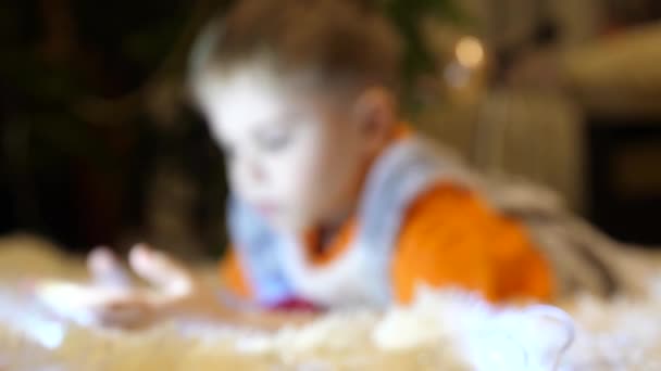 Το παιδί βρίσκεται σε ένα μαλακό κάλυμμα λευκό στο παιδικό δωμάτιο. Αυτός είναι βλέποντας κινούμενα σχέδια στο smartphone. Χριστουγεννιάτικη γιρλάντα. Ευτυχισμένη παιδική ηλικία — Αρχείο Βίντεο