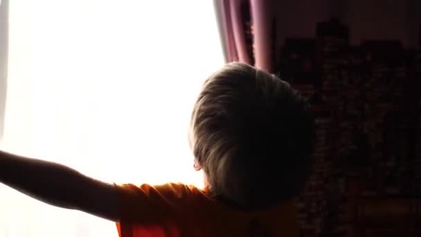 Temprano en la mañana el niño abre las cortinas, los rayos del sol pasan a través de la ventana. amanecer — Vídeo de stock