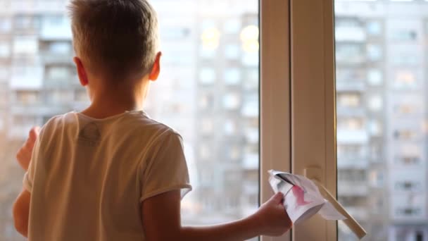 Το παιδί στέκεται στο παράθυρο και κρατά ένα χάρτινο αεροπλάνο. Χειμωνιάτικη μέρα — Αρχείο Βίντεο