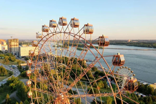 Drone melihat roda Ferris berwarna-warni di taman hiburan di sungai dan lanskap kota. Taman Hiburan dengan roda Ferris besar pada latar belakang dataran tinggi hijau. Tampilan udara Stok Gambar Bebas Royalti