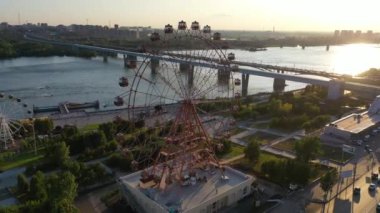Drone nehir ve şehir manzara üzerinde eğlence parkı renkli dönme dolap görünümü. Yeşil dağlık arka plan üzerinde büyük dönme dolap ile eğlence parkı. Havadan görünüm. Gün batımı zamanı