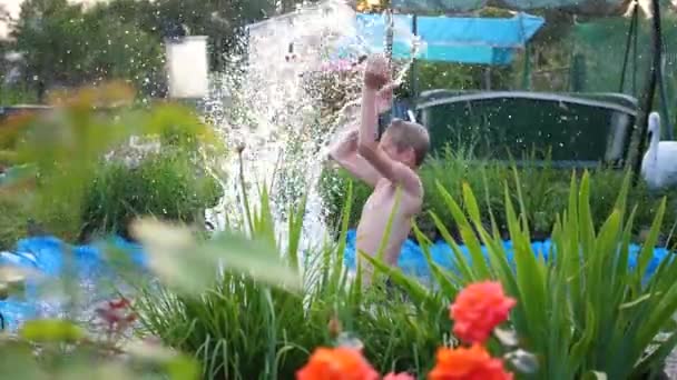 Il nage dans un petit lac par une chaude journée d'été. Le garçon saute dans l'eau, crée des éclaboussures d'eau. Jardin, fleurs et plantes autour du lac. Bonne enfance — Video