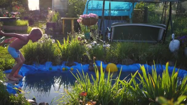 Killen är att simma i en liten sjö på en varm sommardag. Pojken hoppar i vattnet, skapar vattenstänk. Trädgård, blommor och växter runt sjön. Lycklig barndom — Stockvideo