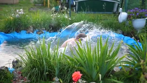 Хлопець плаває в невеликому озері в спекотний літній день. Хлопчик стрибає у воду, створює бризки води. Сад, квіти і рослини навколо озера. Щасливе дитинство — стокове відео