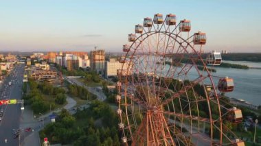 Drone nehir ve şehir manzara üzerinde eğlence parkı renkli dönme dolap görünümü. Yeşil dağlık arka plan üzerinde büyük dönme dolap ile eğlence parkı. Havadan görünüm. Gün batımı zamanı