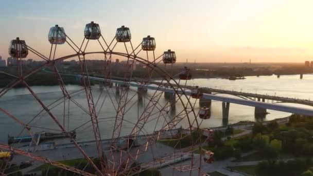 Вид на дрон барвисті колесо огляду в парку атракціонів на річці і на міському ландшафті. Луна-парк з великим колесом огляду на зеленому фоні нагір'я. Вид з повітря. Час заходу сонця — стокове відео