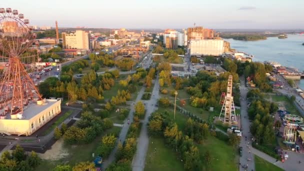 Вид на дрон барвисті колесо огляду в парку атракціонів на річці і на міському ландшафті. Луна-парк з великим колесом огляду на зеленому фоні нагір'я. Вид з повітря. Час заходу сонця — стокове відео