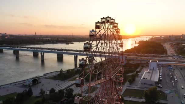 Drone melihat roda Ferris berwarna-warni di taman hiburan di sungai dan lanskap kota. Taman Hiburan dengan roda Ferris besar pada latar belakang dataran tinggi hijau. Pemandangan udara. Waktu matahari terbenam — Stok Video