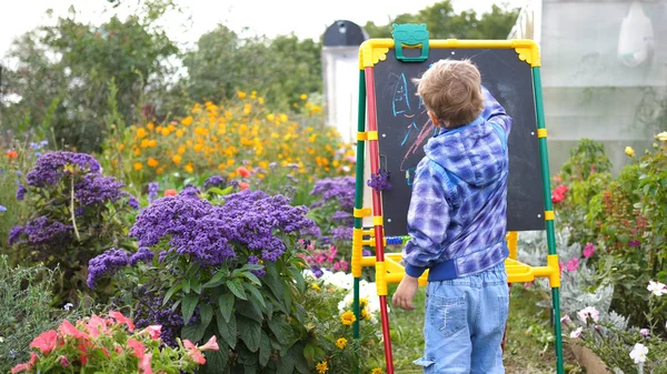 Le jeune artiste s'inspire du conseil d'administration. Un garçon en plein air parmi les fleurs et la verdure dessine des images d'enfants avec de la craie sur le tableau . Photos De Stock Libres De Droits