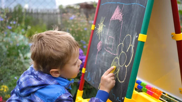 Молодой художник рисует на доске. Мальчик под открытым небом среди цветов и зелени рисует детские рисунки с мелом на доске . — стоковое фото