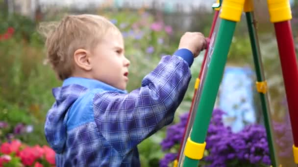 Junge Künstlerin zeichnet auf dem Brett. Ein Junge an der frischen Luft zwischen Blumen und Grün zeichnet Kinderbilder mit Kreide auf die Tafel. — Stockvideo