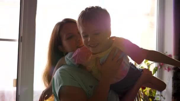 Una madre sostiene a su hijo en sus brazos frente a la ventana. Los rayos del sol pasan a través del vidrio. Buen humor. — Vídeo de stock