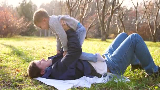 Młody ojciec bawi się z dzieckiem, trzymając je w ramionach. Promienie słońca przenikają przez drzewa. Śmiech i radość całej rodziny. — Wideo stockowe