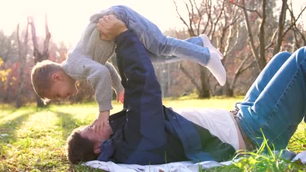 Un padre joven juega con un niño, sosteniéndolo en sus brazos. Los rayos del sol penetran los árboles. Risa y alegría de toda la familia . — Vídeos de Stock