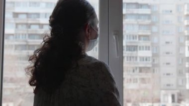 Koruyucu maskeli bir kız kendini izole ederek evde oturuyor. Kız açık pencereden dışarı bakıyor. Sağlık ve can güvenliğinin önlenmesi ve korunması.