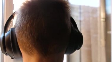 Genç bir adam pencerenin yanında duruyor ve müzik dinlemek için kulaklık takıyor. Gün batımında bulanık arkaplan, gençlik kulaklıkla müzik dinlemekten hoşlanıyor..
