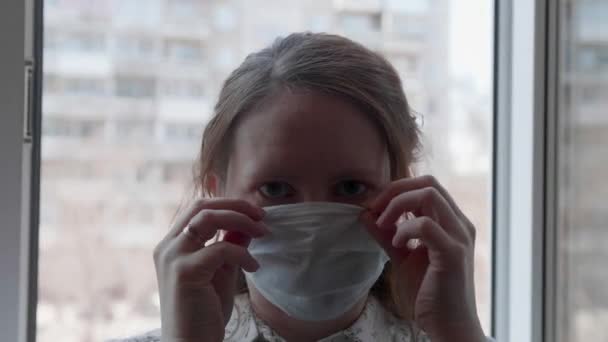 Een meisje met een beschermend masker staat bij een raam in het ziekenhuis. Preventie en bescherming van de gezondheid en de veiligheid van het leven. — Stockvideo