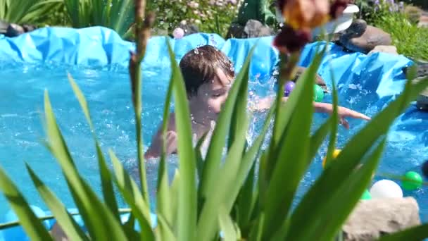 Парень плавает в маленьком бассейне в жаркий летний день. Мальчик прыгает в воду, создавая брызги воды. Сад, цветы и растения вокруг бассейна. Счастливого детства — стоковое видео