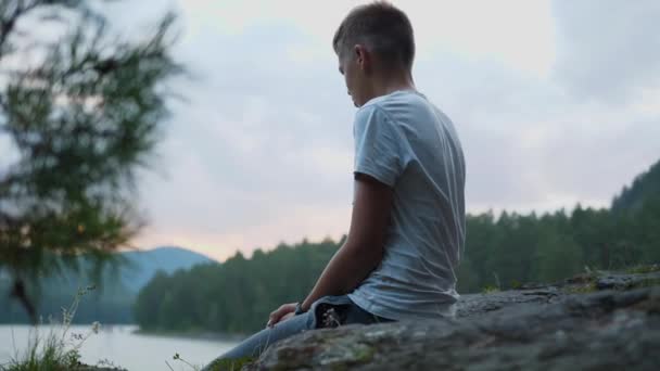 El adolescente se sienta a orillas del río en un acantilado. Tira piedras al agua. — Vídeo de stock