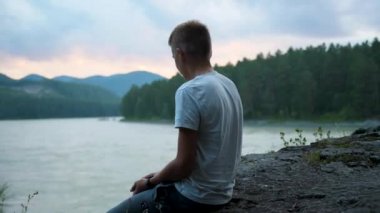 Genç, nehrin kıyısında bir uçurumda oturur. Suya taş atıyor.