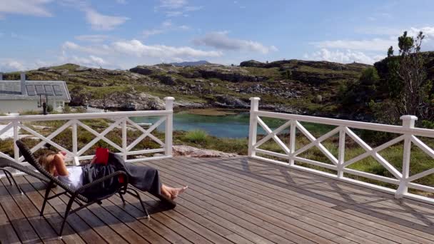 AVEROY, NORWAY, EUROPE - AUGUST 2018: A woman reading a book lying in a trestle on a wooden terrace with a view of the Norwegian fjord in sunny weather, Averoy, Norway — Stock Footage Averoy Norway Europe August 2018 Woman Reading Book Lying Trestle Royalty Free Stock Footage