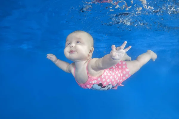 Funny face portrait of little baby girl in the pink dress swim underwater on a blue water background.  Healthy family lifestyle and children water sports activity. Child development, disease prevention
