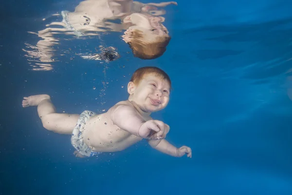 Funny face portrait of baby boy with tongue sticking out swimming and diving underwater with fun in the pool. Healthy family lifestyle and children water sports activity. Child development.